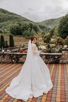 un' dai capelli rossi sposa nel un elegante vestito con lungo maniche e un' bellissimo ornamento nel sua capelli, pose con sua le spalle trasformato per il telecamera su il sfondo di montagne. lungo treno di il vestire. foto