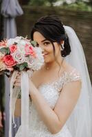 un' brunetta sposa nel un' tiara pose con un' mazzo di bianca e rosa fiori. bellissimo capelli e trucco. primavera nozze foto