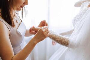 mattina di il sposa. il della sposa domestica di onore aiuta il sposa pizzo su sua vestire, allacciare pulsanti su il vestito o maniche. amiche Aiuto il sposa allacciare sua vestito foto