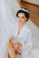 mattina di il sposa prima il nozze. un' bellissimo giovane donna con un' velo e un' bellissimo acconciatura nel un' bianca accappatoio vicino sua nozze vestito su un' manichino. professionale trucco. ultimo preparativi. foto