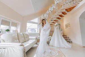 mattina di il sposa prima il nozze. un' bellissimo giovane donna con un' velo e un' bellissimo acconciatura nel un' bianca accappatoio vicino sua nozze vestito su un' manichino. professionale trucco. ultimo preparativi. foto