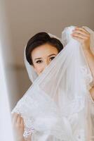 il sposa nel un' seta completo da uomo pose nel sua camera, copertura se stessa con un' velo. mattina di il sposa. preparazione. nozze fotografia foto