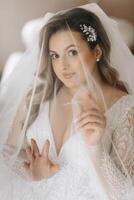 ritratto di un' tenero sposa nel un' nozze vestito e un' bellissimo acconciatura sotto un' velo nel sua camera a partire dal il mattina foto