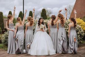 gruppo ritratto di il sposa e damigelle. un' sposa nel un' nozze vestito e damigelle d'onore nel argento vestiti hold elegante mazzi di fiori su loro nozze giorno. foto