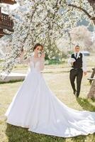 un' bellissimo giovane sposa, nel un' estate parco, passeggiate avanti di sua sposo. bellissimo nozze bianca vestire. passeggiate nel il parco. un' contento e amorevole coppia. foto