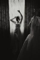 un' donna in piedi nel davanti di un' finestra nel un' condimento toga su un' nozze mattina. nero e bianca foto. foto