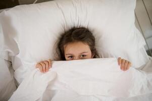 superiore Visualizza di un adorabile bambino ragazza dire bugie su il letto, nascondiglio metà di sua viso sotto bianca lenzuolo, guardare a il telecamera foto