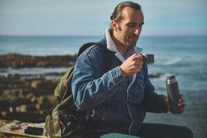 bello viaggiatore uomo detiene un' thermos borraccia e bevande tè o caffè a partire dal inossidabile acciaio tazza, seduta su un' roccia di mare foto