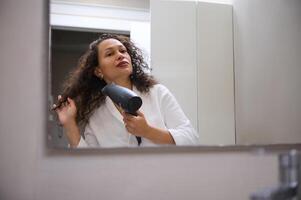 giovane bella donna essiccazione sua lungo bellissimo Riccio capelli con soffio asciugatrice, ammirazione sua riflessione nel il bagno specchio foto