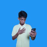 giovane asiatico uomo sorpreso guardare a inteligente Telefono, indossare giallo maglietta, isolato blu sfondo. foto