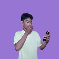 giovane asiatico uomo sorpreso guardare a inteligente Telefono, indossare giallo maglietta, isolato viola sfondo. foto