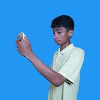 giovane asiatico uomo sorpreso guardare a inteligente Telefono isolato blu sfondo. indossare un' giallo maglietta foto