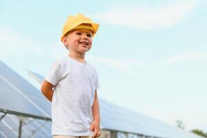 un' contento poco ragazzo nel un' giallo casco è in piedi su un' solare pannello azienda agricola. foto