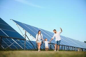 giovane famiglia di tre è accovacciato vicino fotovoltaico solare pannello, poco ragazzo e genitori. moderno famiglia concetto. il concetto di verde energia foto