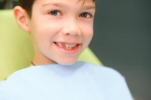 poco ragazzo si siede su dal dentista sedia nel bene umore dopo dentale procedure. giovane paziente con salutare denti foto