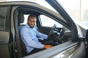 auto proprietario. gioioso afro tipo sorridente, seduta nel nuovo automobile guida a partire dal concessionaria negozio foto