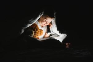 bambino lettura libro nel letto. bambini leggere a notte. poco ragazzo con Fata racconto libri nel Camera da letto . formazione scolastica per giovane bambini. di andare a dormire storia nel il sera. carino ragazzo sotto coperta nel buio camera con lampada foto