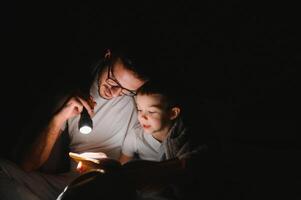 padre e figlio con torcia elettrica lettura libro sotto coperta a casa. foto