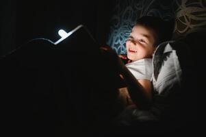 bambino lettura libro nel letto. bambini leggere a notte. poco ragazzo con Fata racconto libri nel Camera da letto . formazione scolastica per giovane bambini. di andare a dormire storia nel il sera. carino ragazzo sotto coperta nel buio camera con lampada foto