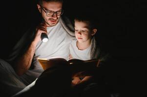 padre e figlio con torcia elettrica lettura libro sotto coperta a casa foto