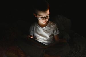 bambino lettura libro nel letto. bambini leggere a notte. poco ragazzo con Fata racconto libri nel Camera da letto . formazione scolastica per giovane bambini. di andare a dormire storia nel il sera. carino ragazzo sotto coperta nel buio camera con lampada foto