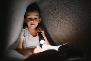 bambino lettura libro nel letto. bambini leggere a notte. poco ragazzo con Fata racconto libri nel Camera da letto . formazione scolastica per giovane bambini. di andare a dormire storia nel il sera. carino ragazzo sotto coperta nel buio camera con lampada foto