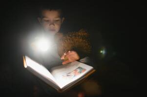 bambino lettura libro nel letto. bambini leggere a notte. poco ragazzo con Fata racconto libri nel Camera da letto . formazione scolastica per giovane bambini. di andare a dormire storia nel il sera. carino ragazzo sotto coperta nel buio camera con lampada foto