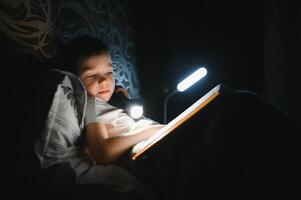 bambino lettura libro nel letto. bambini leggere a notte. poco ragazzo con Fata racconto libri nel Camera da letto . formazione scolastica per giovane bambini. di andare a dormire storia nel il sera. carino ragazzo sotto coperta nel buio camera con lampada foto