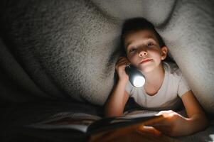 bambino lettura libro nel letto. bambini leggere a notte. poco ragazzo con Fata racconto libri nel Camera da letto . formazione scolastica per giovane bambini. di andare a dormire storia nel il sera. carino ragazzo sotto coperta nel buio camera con lampada foto