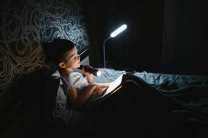 bambino lettura libro nel letto. bambini leggere a notte. poco ragazzo con Fata racconto libri nel Camera da letto . formazione scolastica per giovane bambini. di andare a dormire storia nel il sera. carino ragazzo sotto coperta nel buio camera con lampada foto