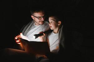 padre e figlio con torcia elettrica lettura libro sotto coperta a casa. foto