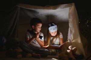 Due bambini con torcia elettrica leggere un' libro sotto un' coperta come un' tenda. foto