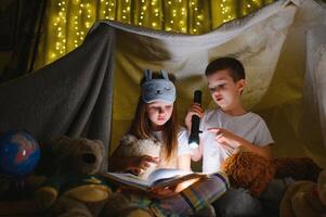 lettura e famiglia Giochi nel figli di tenda. ragazzo e ragazza con libro e torcia elettrica prima andando per letto. foto