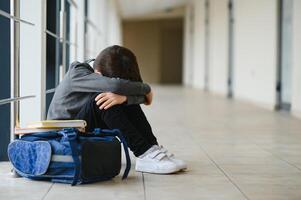 poco ragazzo seduta solo su pavimento dopo sofferenza un atto di bullismo mentre bambini correre nel il sfondo. triste giovane scolaro seduta su corridoio con mani su ginocchia e testa fra il suo gambe foto