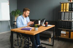 Disabilitato persona nel il sedia a rotelle lavori nel il ufficio a il computer. lui è sorridente e appassionato di il flusso di lavoro. foto