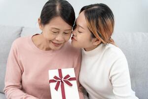 madre giorno, carino asiatico adolescente ragazza abbracciare e baci su il guancia, maturo mezzo età mamma. amore, bacio, cura, contento Sorridi godere famiglia volta. celebrare speciale occasione, contento compleanno, allegro Natale. foto