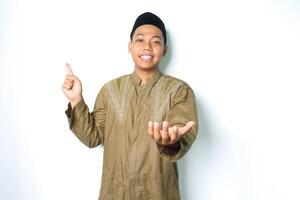 sorridente asiatico musulmano uomo puntamento per sopra e presentazione Aperto palma a telecamera indossare islamico vestito isolato su bianca sfondo foto
