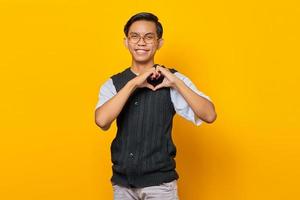 ritratto di allegro giovane uomo asiatico che fa il segno del cuore con le dita su sfondo giallo