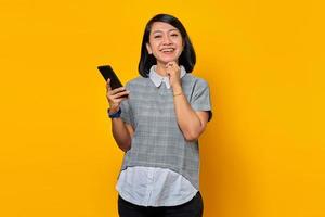 allegra sorridente giovane donna asiatica con il dito sul mento e tenendo il telefono cellulare su sfondo giallo foto