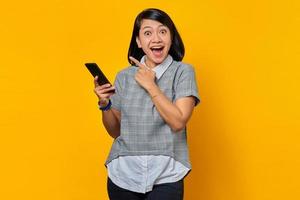 ritratto di una donna asiatica eccitata che tiene in mano il cellulare e punta il dito nello spazio vuoto su sfondo giallo foto