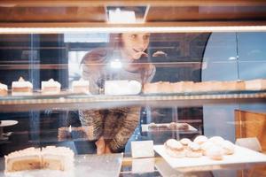 donna sorridente in telecamera attraverso la vetrina con dolci e torte in un moderno bar interno foto