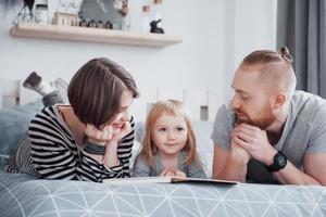 padre, madre e figlia piccola che leggono un libro per bambini su un divano in soggiorno. la grande famiglia felice ha letto un libro interessante in un giorno festivo. i genitori amano i loro figli foto