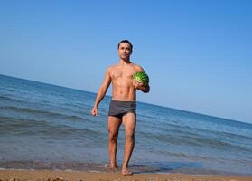 un' sportivo uomo è in piedi su il spiaggia e Tenere un' totale anguria. un' maturo anguria nel il mani di un' uomo foto