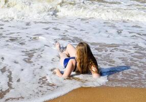 ragazza nel un' blu bagnarsi completo da uomo bugie su il riva del mare e rilassa. onde carezza il ragazze corpo foto