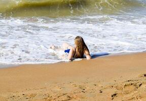 ragazza nel un' blu bagnarsi completo da uomo bugie su il riva del mare e rilassa. onde carezza il ragazze corpo foto