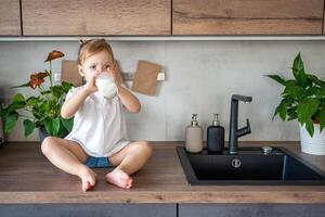 contento bambino ragazza seduta a il tavolo nel il cucina e potabile latte foto