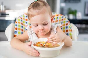 carino bambino ragazza bambino piccolo seduta nel il alto sedia e mangiare sua pranzo la minestra a casa cucina. foto