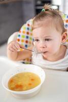 carino bambino ragazza bambino piccolo seduta nel il alto sedia e mangiare sua pranzo la minestra a casa cucina. foto