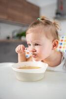 carino bambino ragazza bambino piccolo seduta nel il alto sedia e mangiare sua pranzo la minestra a casa cucina. foto