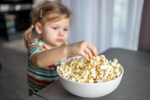 poco ragazza è mangiare Popcorn nel casa cucina. messa a fuoco su mano assunzione Popcorn foto